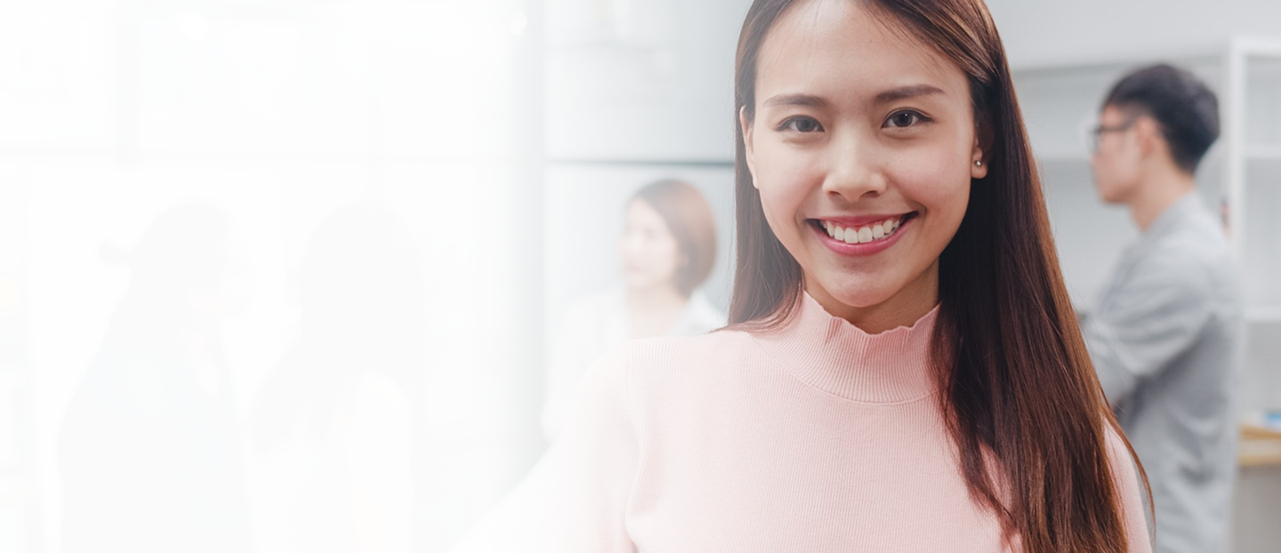 Smiling woman in a pink sweater, representing teamwork and collaboration in a business environment, with colleagues engaged in discussion in the background, highlighting employee benefits and business insurance solutions.