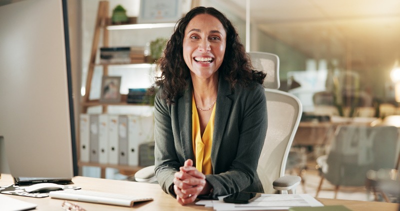 Smiling woman in a professional setting, representing an insurance agent, with documents and a computer on the desk, emphasizing personalized service in health and Medicare insurance planning.