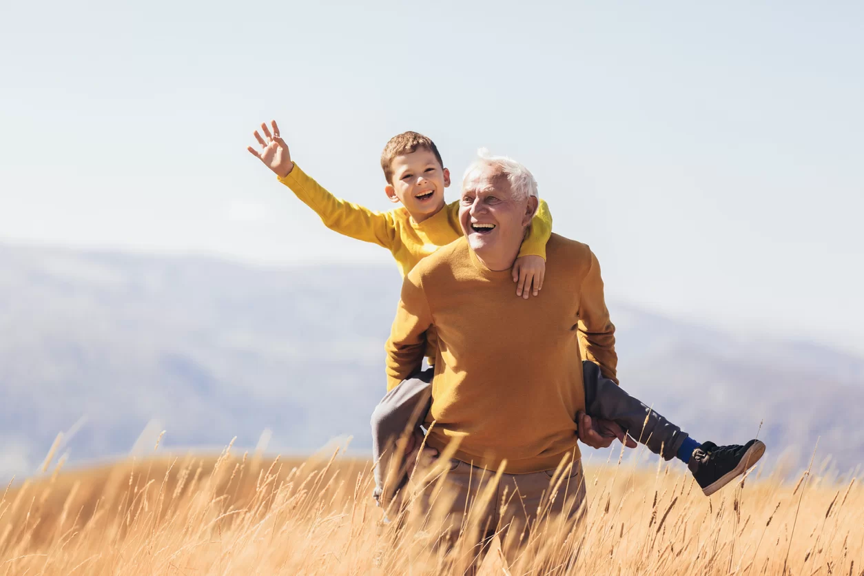 Grandfather carrying grandson on shoulders, both smiling in a sunny field, symbolizing family bonds and joyful moments related to senior life insurance.
