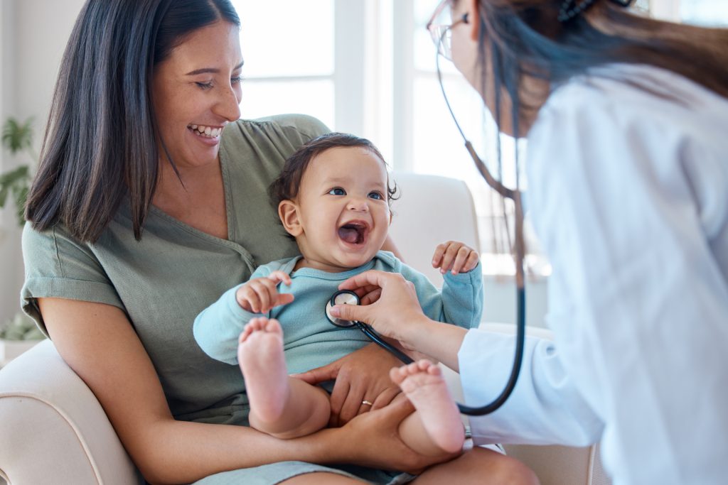 Mother smiling while holding a happy baby during a pediatric check-up with a doctor using a stethoscope, emphasizing family health and healthcare plans.