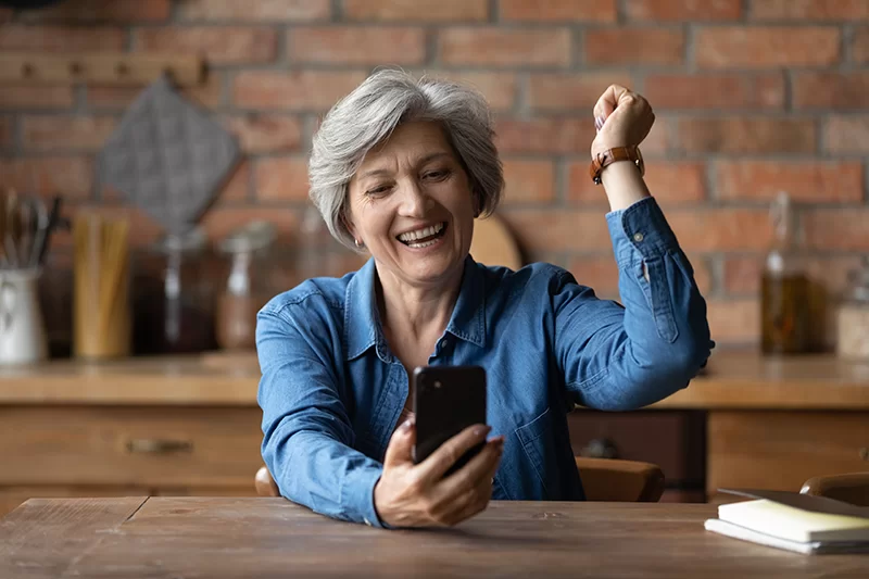 Senior woman smiling and celebrating while using a smartphone, representing engagement with financial planning and insurance information.