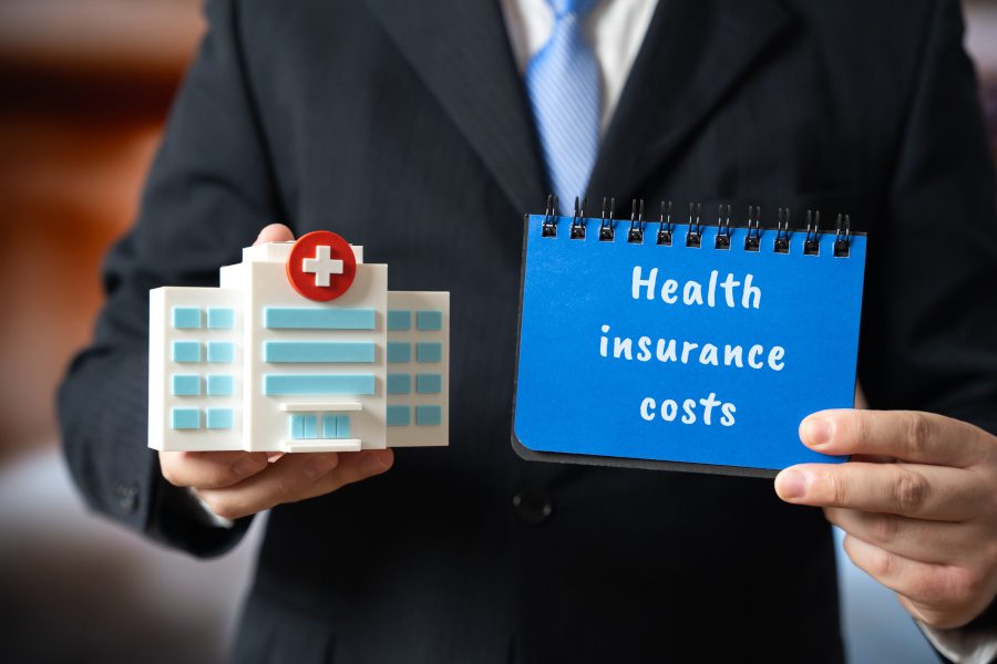 man holding a model of a hospital and a notebook saying, "Health insurance costs"