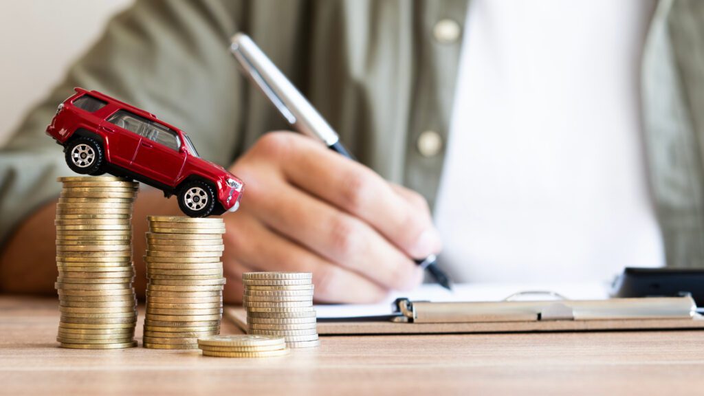 Red toy car on stacked coins with person writing notes, symbolizing auto insurance costs and financial planning.