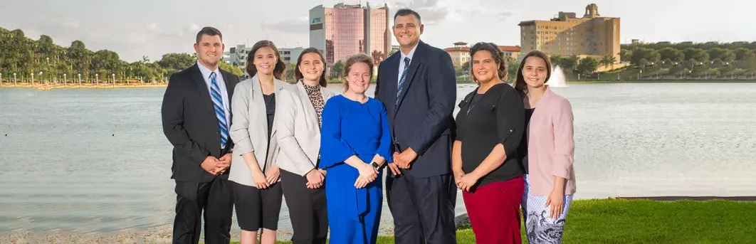 A diverse group of seven professionals posing together by a lake with buildings in the background.